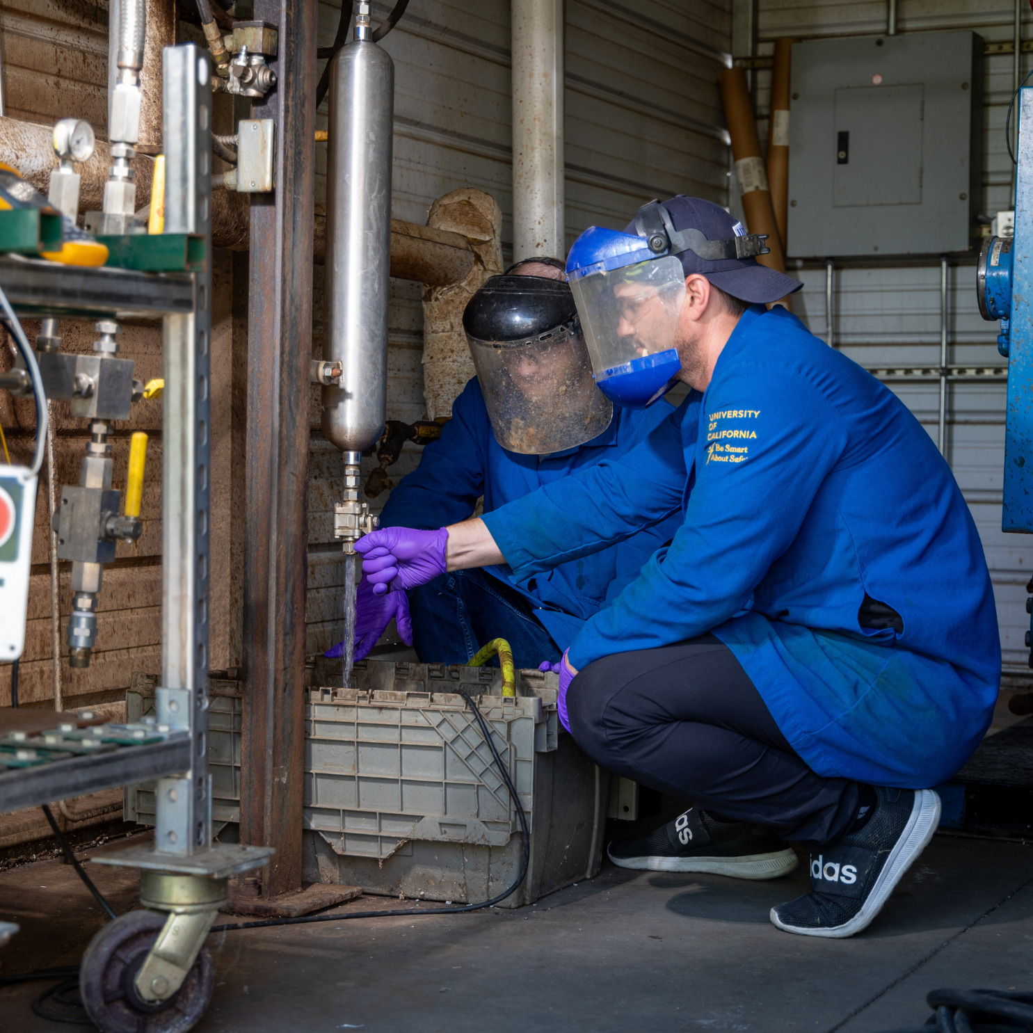 Two researchers wearing face shields, blue lab coats, and protective gloves work together to operate laboratory equipment during an experiment in an industrial research facility.