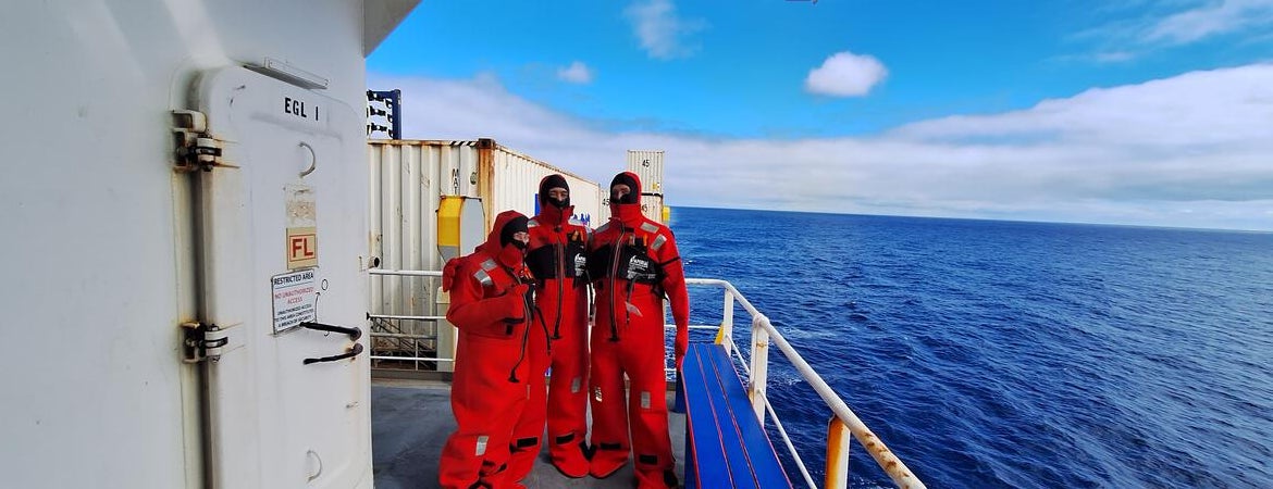 Three researchers wearing bright red immersion suits stand on the deck of a ship, with the open ocean and partly cloudy blue sky in the background. The group is positioned near a white metal door and railing, with containers and equipment visible on the deck.