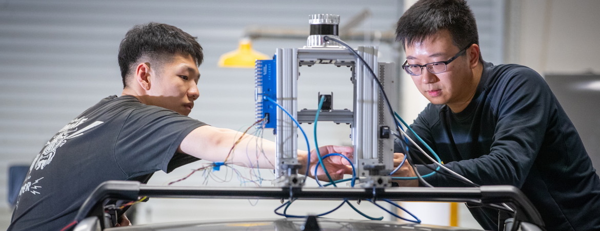 Two researchers adjust a sensor-equipped apparatus mounted on top of a vehicle, working with exposed wiring and instrumentation in a laboratory setting.