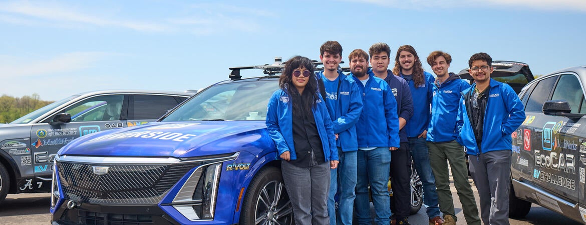Seven members of the UC Riverside EcoCAR team stand beside R'VOLT, their blue Cadillac LYRIQ competition vehicle, at the General Motors Milford Proving Ground during the final EcoCAR EV Challenge in Michigan. Team members wear matching blue UCR EcoCAR jackets, with two other competition vehicles visible in the background under a clear sky.
