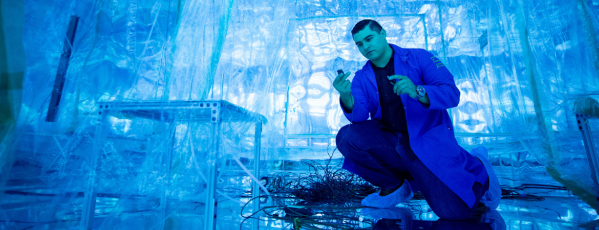 Researcher wearing a blue lab coat kneels inside the blue-lit Advanced Plant Laboratory (APL), inspecting sensors and wiring within a controlled-environment research chamber.