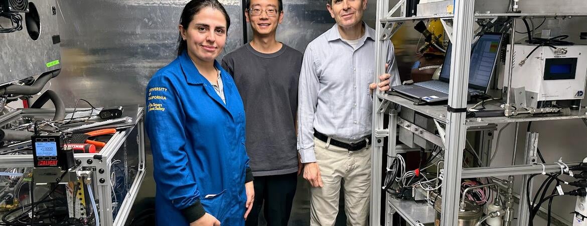 A front-view, full-body shot of three UCR CE-CERT researchers standing inside a mobile lab truck. From left to right: graduate student Maryam Yousefi, in a blue university jacket; graduate student Minghao Han, in a gray t-shirt and glasses; and CE-CERT Director Dr. Don Collins, in a collared shirt. They are all looking at the camera and smiling faintly. The mobile lab has shiny, corrugated metal walls and is filled with intricate scientific equipment on metal racks, with many wires visible.