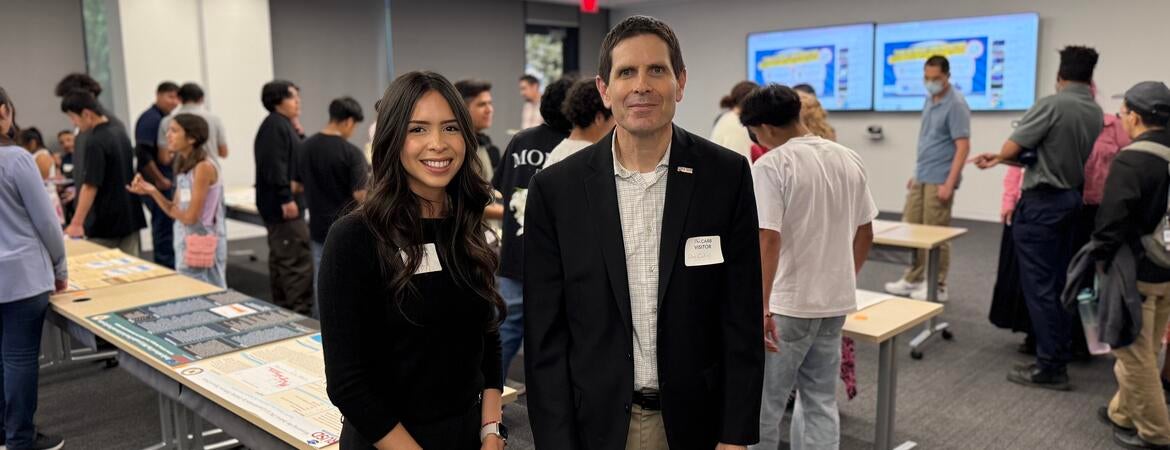 Kiara Salgado, a young woman with long dark hair wearing a black outfit, stands next to Don Collins, who is wearing a black blazer and khaki pants. They are both smiling at the camera in a classroom filled with students and poster presentations displayed on tables in the background.
