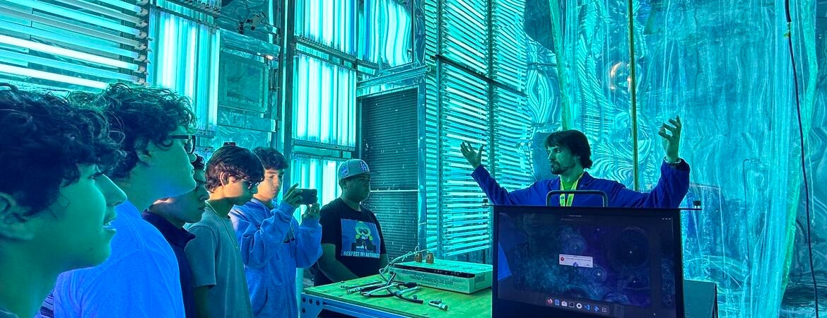 A group of five high school students on a STEPCon (Science and Technology Education Partnership Conference) tour are gathered in the world's largest indoor atmospheric processes chamber at the UC Riverside CE-CERT Atmospheric Processes Laboratory. The students are intently focused on a researcher in a blue jacket, who is enthusiastically explaining a concept with his arms raised, standing near a laptop. The massive chamber is dramatically lit by arrays of intense cyan/blue UV lights