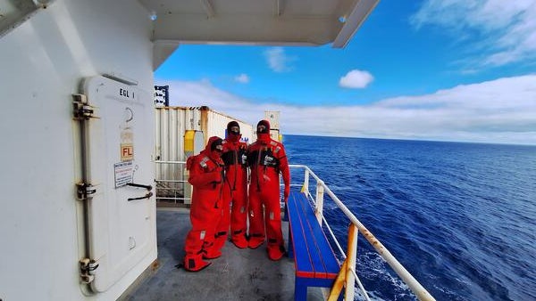 Three researchers wearing bright red immersion suits stand on the deck of a ship, with the open ocean and partly cloudy blue sky in the background. The group is positioned near a white metal door and railing, with containers and equipment visible on the deck.