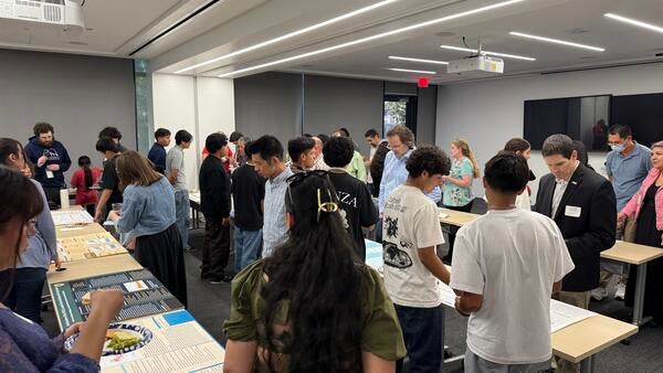 A classroom filled with students and adults viewing air quality research posters displayed on long tables. Attendees are reading, discussing, and pointing at various projects. The room is brightly lit with digital screens and projectors at the front. Don Collins is visible on the right side of the image, reviewing a student’s project.