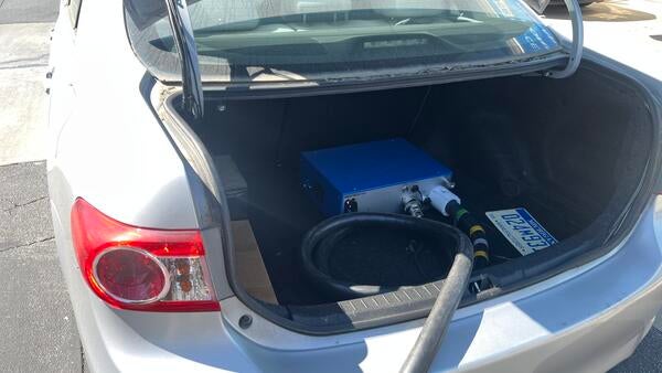Rear view of a silver Toyota Corolla parked in a lot with a black emissions sampling hose extending from the trunk, indicating mobile emissions testing in progress.