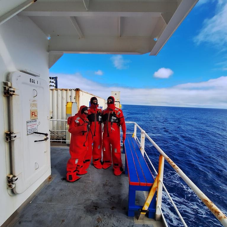 Three researchers wearing bright red immersion suits stand on the deck of a ship, with the open ocean and partly cloudy blue sky in the background. The group is positioned near a white metal door and railing, with containers and equipment visible on the deck.