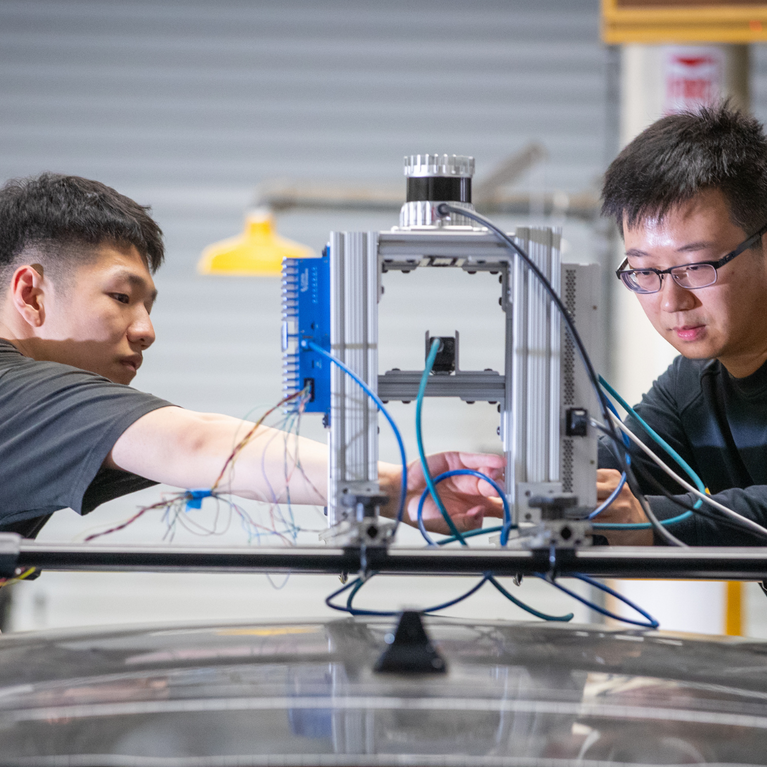 Two researchers adjust a sensor-equipped apparatus mounted on top of a vehicle, working with exposed wiring and instrumentation in a laboratory setting.