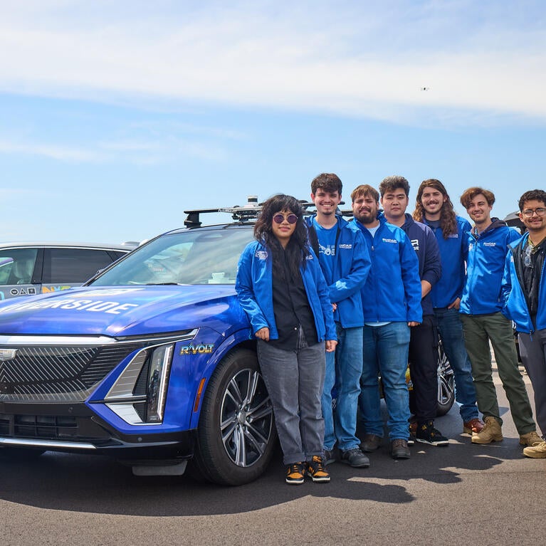 Seven members of the UC Riverside EcoCAR team stand beside R'VOLT, their blue Cadillac LYRIQ competition vehicle, at the General Motors Milford Proving Ground during the final EcoCAR EV Challenge in Michigan. Team members wear matching blue UCR EcoCAR jackets, with two other competition vehicles visible in the background under a clear sky.