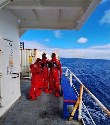 Three researchers wearing bright red immersion suits stand on the deck of a ship, with the open ocean and partly cloudy blue sky in the background. The group is positioned near a white metal door and railing, with containers and equipment visible on the deck.