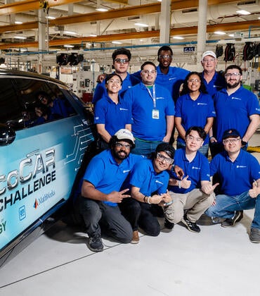 The UCR EcoCAR team gathers for a group photo next to their Cadillac LYRIQ in the workshop, all wearing matching blue team polos and smiling for the camera.