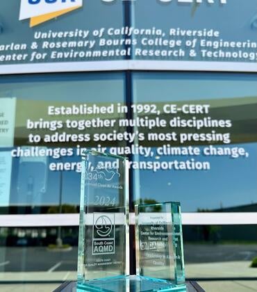 A glass award displayed in front of the University of California, Riverside's CE-CERT building, reflecting the text "University of California, Riverside Bourns College of Engineering Center for Environmental Research & Technology." The award is from the South Coast Air Quality Management District, recognizing the center for "Leadership in Air Quality." The background text highlights CE-CERT's mission to address air quality, climate change, energy, and transportation challenges