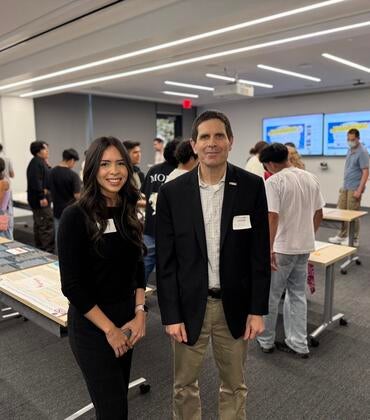 Kiara Salgado, a young woman with long dark hair wearing a black outfit, stands next to Don Collins, who is wearing a black blazer and khaki pants. They are both smiling at the camera in a classroom filled with students and poster presentations displayed on tables in the background.