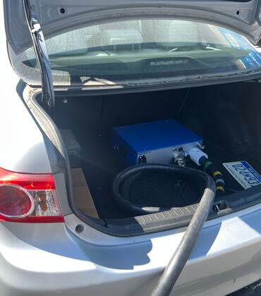 pen trunk of a silver Toyota Corolla showing a blue portable emissions analyzer connected to a black hose, used for measuring ammonia emissions during on-road testing.