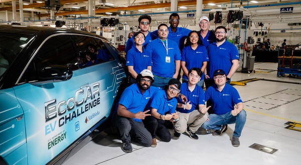 The UCR EcoCAR team gathers for a group photo next to their Cadillac LYRIQ in the workshop, all wearing matching blue team polos and smiling for the camera.