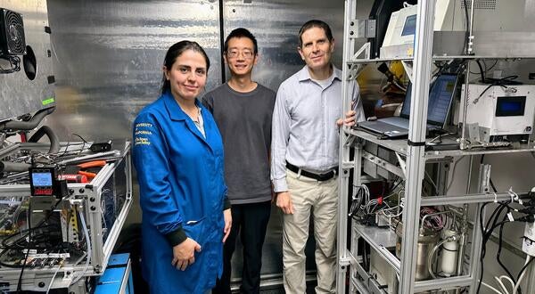 A front-view, full-body shot of three UCR CE-CERT researchers standing inside a mobile lab truck. From left to right: graduate student Maryam Yousefi, in a blue university jacket; graduate student Minghao Han, in a gray t-shirt and glasses; and CE-CERT Director Dr. Don Collins, in a collared shirt. They are all looking at the camera and smiling faintly. The mobile lab has shiny, corrugated metal walls and is filled with intricate scientific equipment on metal racks, with many wires visible.