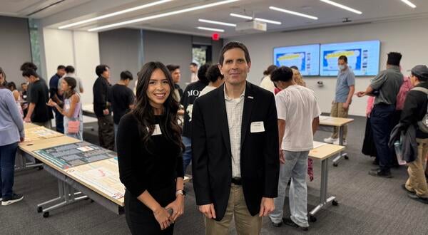 Kiara Salgado, a young woman with long dark hair wearing a black outfit, stands next to Don Collins, who is wearing a black blazer and khaki pants. They are both smiling at the camera in a classroom filled with students and poster presentations displayed on tables in the background.