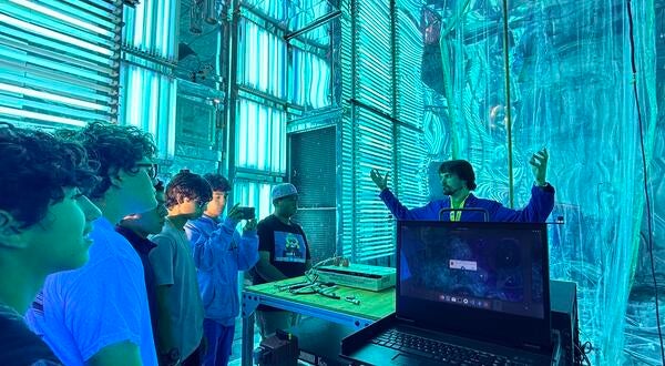 A group of five high school students on a STEPCon (Science and Technology Education Partnership Conference) tour are gathered in the world's largest indoor atmospheric processes chamber at the UC Riverside CE-CERT Atmospheric Processes Laboratory. The students are intently focused on a researcher in a blue jacket, who is enthusiastically explaining a concept with his arms raised, standing near a laptop. The massive chamber is dramatically lit by arrays of intense cyan/blue UV lights