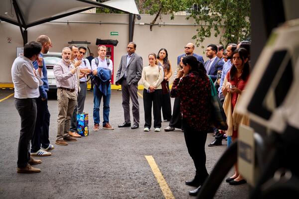 A group of ZEV Working Group members gathers in a semicircle during a technical site tour of electric mobility infrastructure in Mexico City, with a speaker addressing the group while others listen attentively.