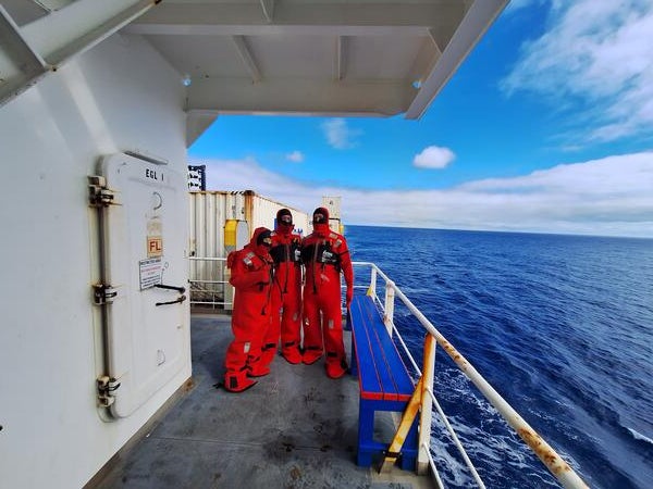 Three researchers wearing bright red immersion suits stand on the deck of a ship, with the open ocean and partly cloudy blue sky in the background. The group is positioned near a white metal door and railing, with containers and equipment visible on the deck.
