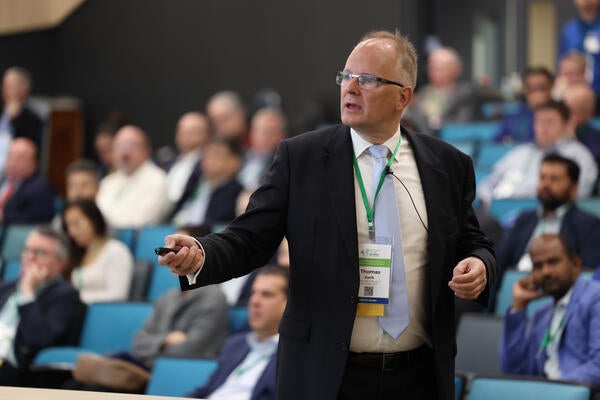 Professor Thomas Koch, keynote speaker from Karlsruhe Institute of Technology and CEO of Allianz Wasserstoffmotor, speaks to attendees during the Inaugural 2025 Hydrogen Engine Conference. He is holding a presentation clicker, wearing a suit and conference badge, with a full audience seated in the background.