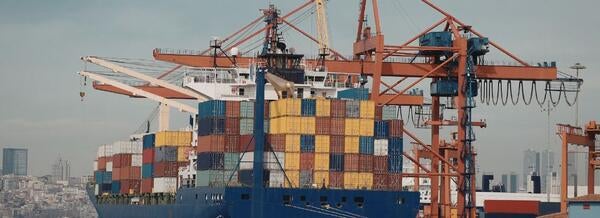 Container ship docked at a port with stacked shipping containers and large cranes loading cargo in the background.