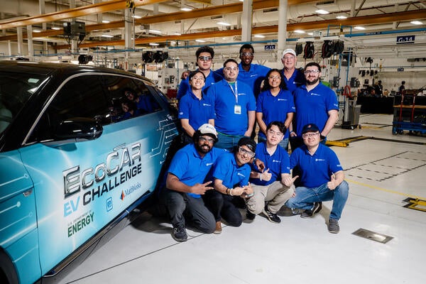 The UCR EcoCAR team gathers for a group photo next to their Cadillac LYRIQ in the workshop, all wearing matching blue team polos and smiling for the camera.