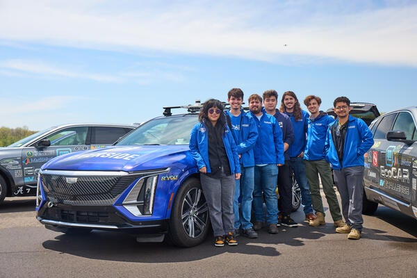Seven members of the UC Riverside EcoCAR team stand beside R'VOLT, their blue Cadillac LYRIQ competition vehicle, at the General Motors Milford Proving Ground during the final EcoCAR EV Challenge in Michigan. Team members wear matching blue UCR EcoCAR jackets, with two other competition vehicles visible in the background under a clear sky.