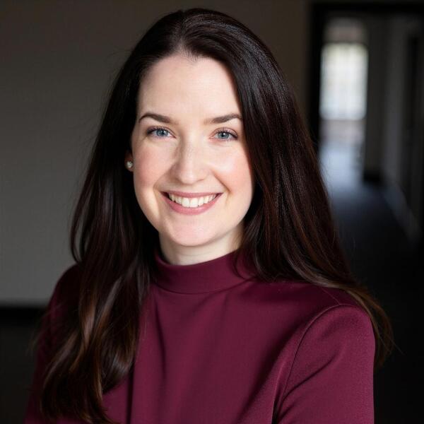 A professional headshot of Stephanie Severance, a woman with long, dark brown hair and blue eyes, smiling warmly. She is wearing a high-necked, maroon top and small stud earrings. The background is a softly blurred indoor setting.