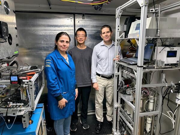 A front-view, full-body shot of three UCR CE-CERT researchers standing inside a mobile lab truck. From left to right: graduate student Maryam Yousefi, in a blue university jacket; graduate student Minghao Han, in a gray t-shirt and glasses; and CE-CERT Director Dr. Don Collins, in a collared shirt. They are all looking at the camera and smiling faintly. The mobile lab has shiny, corrugated metal walls and is filled with intricate scientific equipment on metal racks, with many wires visible.