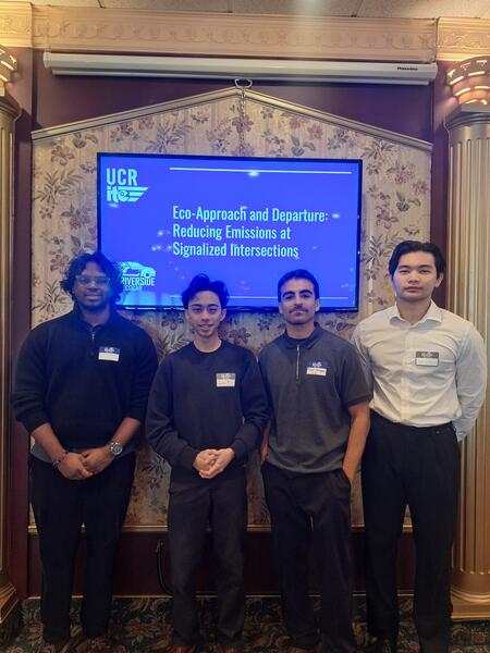 Four students standing in front of a presentation screen displaying the title 'Eco-Approach and Departure: Reducing Emissions at Signalized Intersections' at a UCR event. All are wearing name tags and business casual attire.
