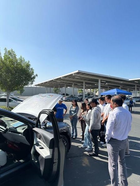The UCR EcoCAR team presentation during CE-CERT's Open House. A group of approximately 10 high school or university students are gathered outdoors on a sunny day in a parking lot. They are listening to a man in a blue shirt standing over the open hood of a dark car, which has its driver's side door open. The EcoCAR project information is visible on the hood. A large solar panel structure is visible in the background.