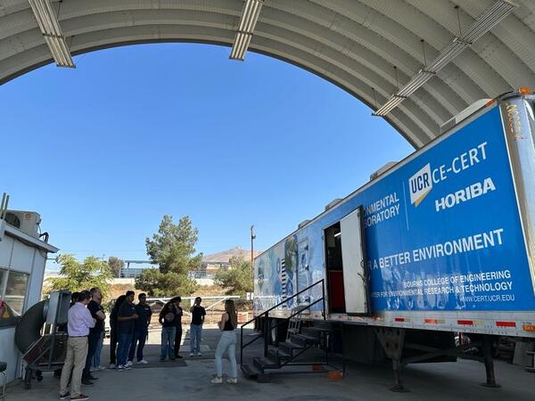 A group of students and staff are gathered under the metal bay of the heavy-duty chassis dynamometer at CE-CERT, listening to a presentation. The bay houses the large, blue Mobile Emissions Laboratory (MEL) trailer, which is used for real-time emissions testing. The trailer is branded with 'UCR CE-CERT HORIBA' and 'FOR A BETTER ENVIRONMENT'.