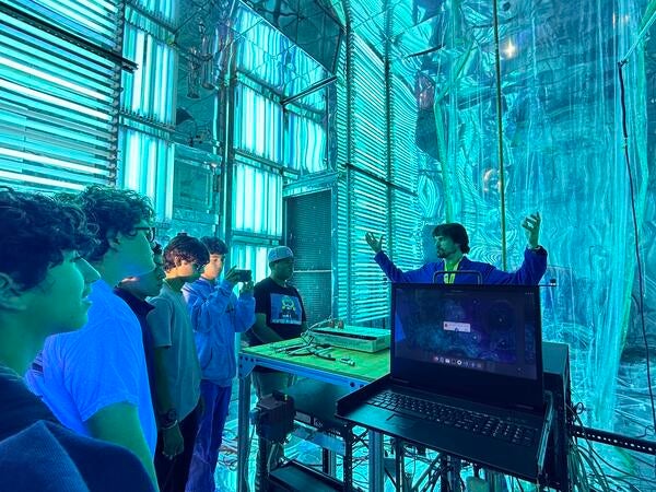 A group of five high school students on a STEPCon (Science and Technology Education Partnership Conference) tour are gathered in the world's largest indoor atmospheric processes chamber at the UC Riverside CE-CERT Atmospheric Processes Laboratory. The students are intently focused on a researcher in a blue jacket, who is enthusiastically explaining a concept with his arms raised, standing near a laptop. The massive chamber is dramatically lit by arrays of intense cyan/blue UV lights