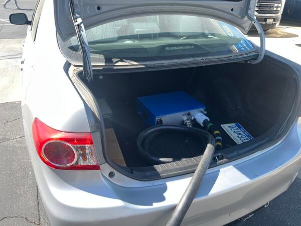 Rear view of a silver Toyota Corolla parked in a lot with a black emissions sampling hose extending from the trunk, indicating mobile emissions testing in progress.