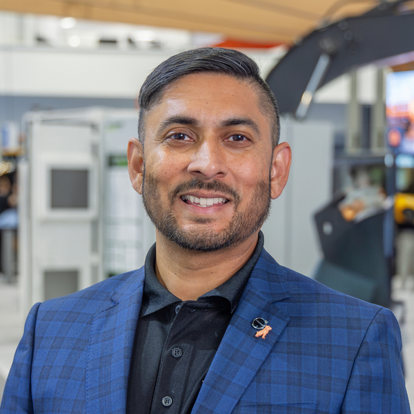 a man wearing a blue checkered blazer and black collared shirt smiles at the camera in an indoor professional setting with blurred equipment and displays in the background.
