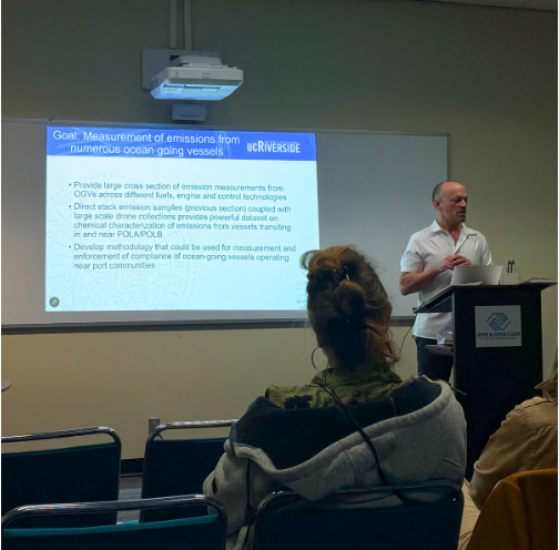 A CE-CERT researcher presents project goals during a community meeting in San Pedro while residents listen from the audience. A projected slide outlines plans to measure emissions from oceangoing vessels and evaluate their impacts on air quality in nearby port communities.