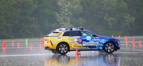 R'VOLT, the UC Riverside EcoCAR team's reengineered Cadillac LYRIQ, drives through a rain-soaked test course lined with orange traffic cones during dynamic vehicle testing at the final EcoCAR EV Challenge in Michigan. Water sprays from the tires as the vehicle completes a handling event.