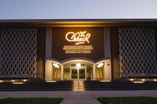 cheech The Cheech Marin Center for Chicano Art & Culture in Riverside, California, shown at dusk with its illuminated “The Cheech” sign glowing above the entrance. Modern latticework panels flank the building’s front, and wide steps with accent lighting lead up to the glass doors.